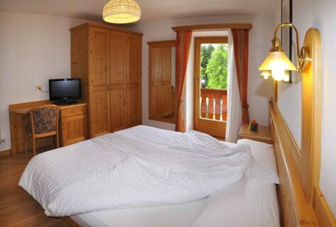 Světle zař&iacute;zen&aacute; ložnice v hotelu Albergo Nido Verde s manželskou postel&iacute;, dřevěn&yacute;m n&aacute;bytkem, TV, &scaron;atn&iacute; skř&iacute;n&iacute; a balkonem s v&yacute;hledem do zeleně.
