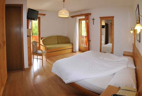 Světl&yacute; a &uacute;tuln&yacute; pokoj v hotelu Albergo Nido Verde s manželskou postel&iacute;, pohovkou, dřevěn&yacute;m n&aacute;bytkem a balkonem s v&yacute;hledem do zeleně.