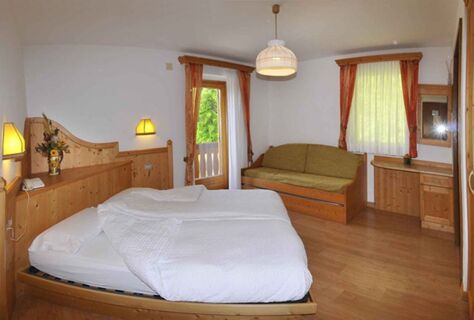 &Uacute;tuln&yacute; hotelov&yacute; pokoj s dřevěn&yacute;m n&aacute;bytkem, manželskou postel&iacute; a pohovkou v hotelu Albergo Nido Verde s v&yacute;hledem do zelen&eacute; př&iacute;rody z okna a balkonu.
