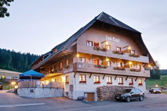 Фасад традиционного отеля Gasthaus Zum Engel со сваями из дерева, балконами с цветами и открытой площадкой с автомобилями в вечернее время.