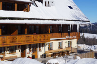 Фасад отеля Gasthaus Zum Engel с заснеженной крышей, большими сосульками и деревянной верандой зимой под ясным небом.