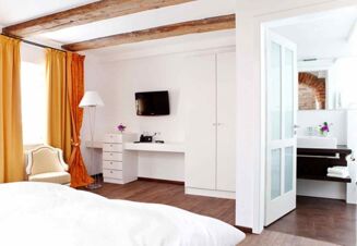 A hotel room at Brauereigasthof und Hotel Kapplerbr&auml;u featuring white walls, wooden ceiling beams, mustard curtains, a TV, wardrobe, and a view into the bathroom with flowers