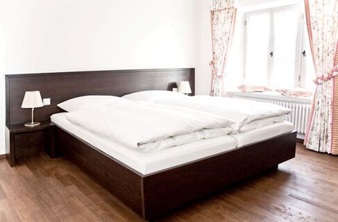 A clean double bed with white bedding and pillows in a bright room with wooden floor and floral curtains at Brauereigasthof und Hotel Kapplerbr&auml;u