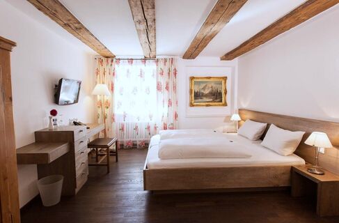 Bedroom with double bed, wooden ceiling beams, floral curtains, wall painting, and desk at Brauereigasthof und Hotel Kapplerbr&auml;u