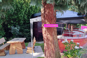 Wooden picnic table with benches beside a tree and a covered red boat near Hotel Columbus & Glamping greenery