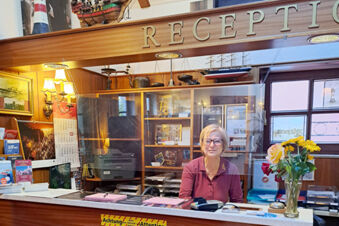 A receptionist sitting behind a counter with a transparent screen at Hotel Columbus & Glamping reception area with flowers and brochures