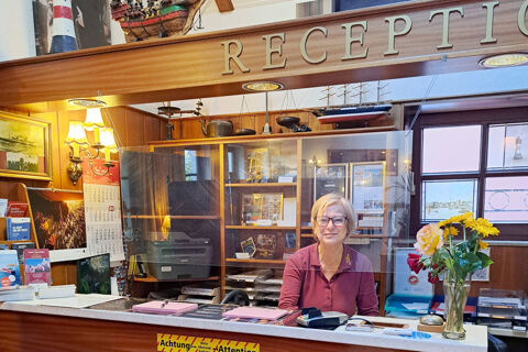 A receptionist sitting behind a counter with a transparent screen at Hotel Columbus & Glamping reception area with flowers and brochures