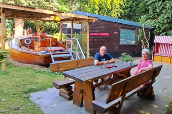 A man and woman sitting at a wooden picnic table outside near a small wooden boat and cabin at Hotel Columbus & Glamping