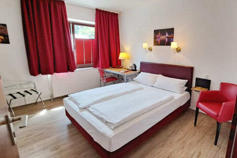 A clean hotel room at Hotel Columbus & Glamping with a double bed, red curtains, a desk with a lamp, and a red armchair