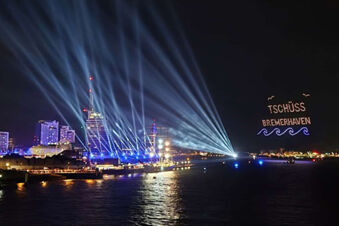 Night view of a waterfront city with light beams and illuminated text "TSCH&Uuml;SS BREMERHAVEN" over water near Hotel Columbus & Glamping