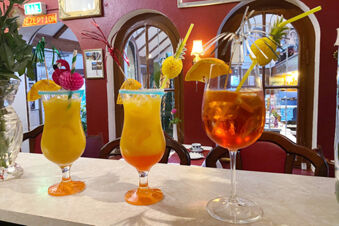 Three colorful cocktails with fruit garnishes and straws on a marble counter at Hotel Columbus & Glamping interior bar area