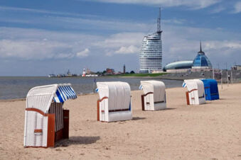 Five white and blue striped beach chairs on sand near water with modern buildings in the background at Hotel Columbus & Glamping