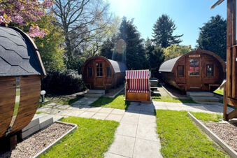 Three wooden barrel-shaped cabins with names, a striped lounge chair, and surrounding grass and trees at Hotel Columbus & Glamping