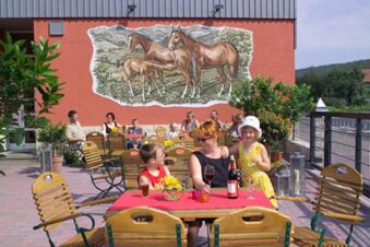People sitting at wooden tables with drinks outdoors under a mural of three horses at Rh&ouml;ngasthof Zur Linde & Ferien-Appartements Rh&ouml;nsicht
