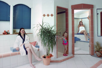 Woman in a white bathrobe sitting on a tiled bench, child in a swimsuit in a shower, and woman in a towel at Rh&ouml;ngasthof Zur Linde & Ferien-Appartements Rh&ouml;nsicht