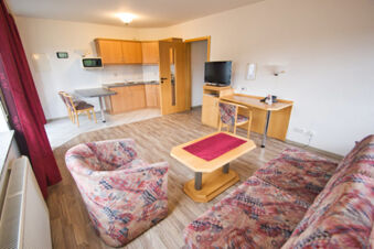 Living area with patterned sofa and armchair, wooden coffee table, kitchen with cabinets, and TV at Rh&ouml;ngasthof Zur Linde & Ferien-Appartements Rh&ouml;nsicht