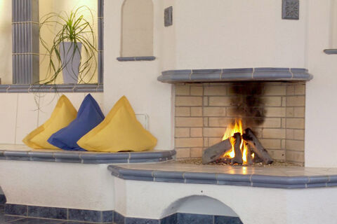 Fireplace with burning logs and yellow and blue cushions on a built-in tiled bench at Rh&ouml;ngasthof Zur Linde & Ferien-Appartements Rh&ouml;nsicht