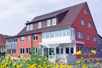 A red and gray multi-story building with many windows and white entrance labeled Gasthof Zur Linde and yellow flowers in front for Rh&ouml;ngasthof Zur Linde & Ferien-Appartements Rh&ouml;nsicht