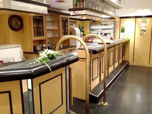 Elegante Bar mit Holzverkleidung und Glasregalen in K&ouml;hncke's Hotel, ausgestattet mit Theke und Barhockern.