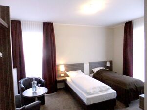 Ein schlicht eingerichtetes Doppelzimmer mit zwei Betten, Sessel und Tisch in K&ouml;hncke's Hotel mit lichtdurchfluteten Fenstern.
