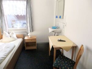 Kleines Zimmer mit Einzelbett, Nachttisch, Tisch mit Stuhl und Waschbecken im Zimmer bei K&ouml;hncke's Hotel, schlicht eingerichtet.
