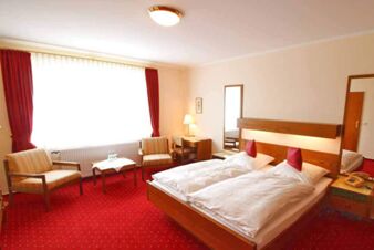 Helles Hotelzimmer mit Doppelbett, roten Teppichen und Sitzbereich im Landhotel Helms mit schlichtem Holzmobiliar.