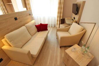 Hell eingerichtetes Zimmer im Landhotel Helms mit Sofa, Sessel, Holzfu&szlig;boden und Fenster mit Vorh&auml;ngen sowie einem TV an der Wand.