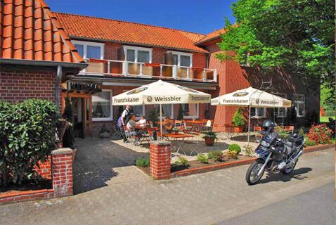 Au&szlig;enbereich des Landhotel Helms mit roten Ziegelgeb&auml;uden, Tischen unter Sonnenschirmen und einem geparkten Motorrad bei Sonnenschein.