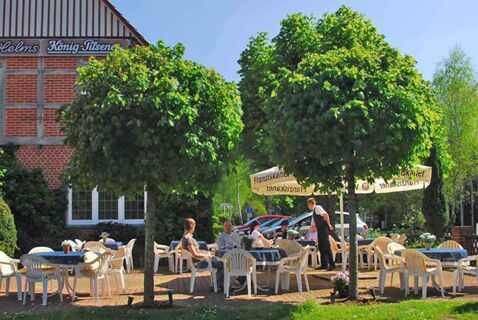 Im Au&szlig;enbereich des Landhotel Helms sitzen G&auml;ste an Tischen unter Sonnenschirmen und B&auml;umen bei sonnigem Wetter.