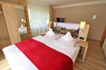 Helles Hotelzimmer mit Doppelbett, roter Tagesdecke und Naturmotiv am Kopfteil im Landhotel Helms mit hellem Holzmobiliar.