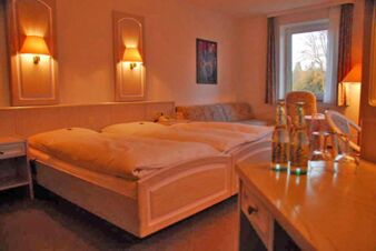 Gem&uuml;tliches Doppelzimmer im Landhotel Helms mit warmem Licht, zwei Betten, Sofa, Holzm&ouml;beln und Fenster mit Blick ins Gr&uuml;ne.