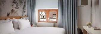 Modern eingerichtetes Hotelzimmer mit wei&szlig;er Bettw&auml;sche und Fenster mit Blick auf historische Geb&auml;ude im NH Collection Hotel Heidelberg.