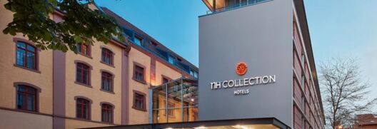 Ansicht der modernen Fassade des NH Collection Hotel Heidelberg mit beleuchtetem Logo und angrenzendem historischen Geb&auml;udeteil bei Abendlicht.
