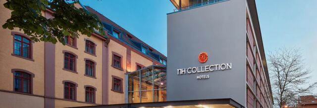 Ansicht der modernen Fassade des NH Collection Hotel Heidelberg mit beleuchtetem Logo und angrenzendem historischen Geb&auml;udeteil bei Abendlicht.