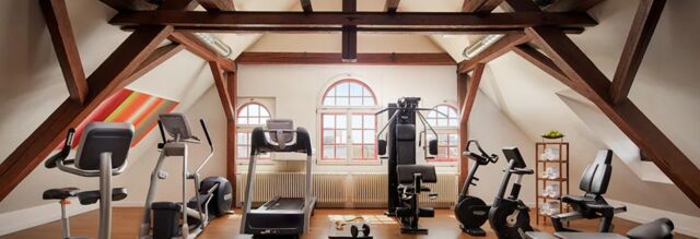 Fitnessbereich mit modernen Trainingsger&auml;ten im Dachgeschoss des NH Collection Hotel Heidelberg mit gro&szlig;en Fenstern und Holzbalken.