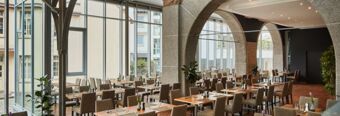 Helles und modernes Restaurant mit Holztischen und beigen St&uuml;hlen im NH Collection Hotel Heidelberg mit gro&szlig;en Glasfenstern und Steinb&ouml;gen.