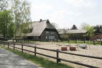 Das Foto zeigt eine traditionelle Reetdach-Fachwerkhaus-Fassade mit einem umz&auml;unten Sandreitplatz bei Ferienhof Klaucke im l&auml;ndlichen Umfeld.