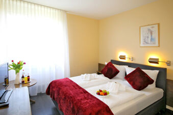 Helles und gem&uuml;tliches Doppelzimmer im Hotel Kick mit roten Kissen, Tagesdecke, Fernseher und einem kleinen Obstteller auf dem Bett.