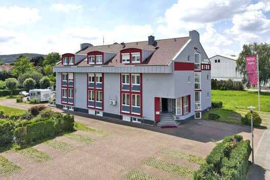 Das moderne Geb&auml;ude des Hotel Kick mit roten und wei&szlig;en Akzenten, einem gepflasterten Parkplatz und gr&uuml;ner Umgebung bei Tageslicht.