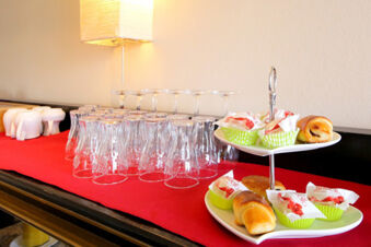 Ein Buffet mit Gl&auml;sern, Kaffetassen und Geb&auml;ck auf einem roten Tuch, arrangiert im Hotel Kick f&uuml;r G&auml;ste.