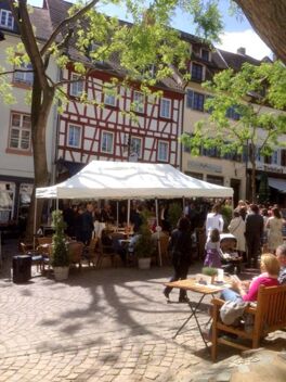 Au&szlig;enansicht eines belebten Marktplatzes mit G&auml;sten unter einem wei&szlig;en Zelt und Sitzgelegenheiten beim Marktplatzhotel u. Bistrorant Tafelspitz.