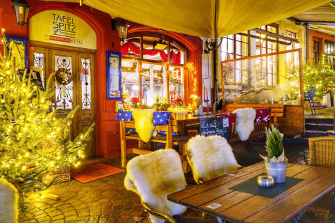 Au&szlig;enansicht des festlich beleuchteten Marktplatzhotel u. Bistrorant Tafelspitz mit warm dekorierter Terrasse und gem&uuml;tlichen Sitzgelegenheiten unter einem gelben Sonnenschirm.