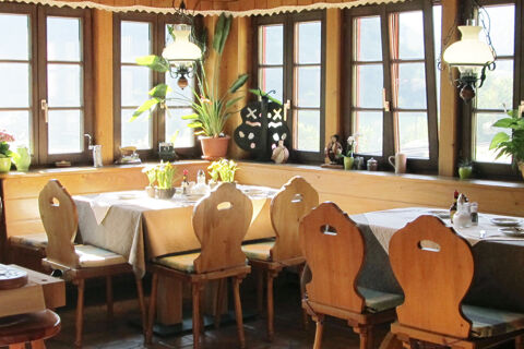 Gem&uuml;tlicher Restaurantbereich mit Holzst&uuml;hlen und Tischen vor gro&szlig;en Fenstern im Bauernhof W&auml;ldebauernhof, hell und einladend gestaltet.