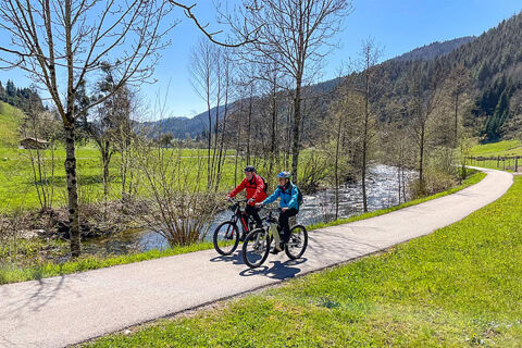 Zwei Radfahrer fahren einen Weg entlang eines Bachs in der gr&uuml;nen Natur, umgeben von Bergen beim Bauernhof W&auml;ldebauernhof.