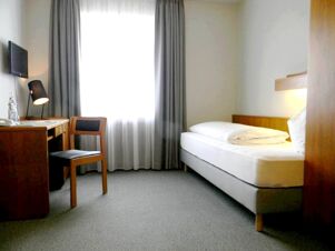 Helles Einzelzimmer mit schlichtem Design im Hotel Landgut Burg, ausgestattet mit Bett, Schreibtisch und Stuhl vor einem Fenster mit Vorh&auml;ngen.
