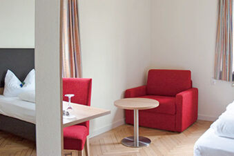 Ein gem&uuml;tliches Hotelzimmer im Hotel Landgut Burg mit rotem Sessel, kleinem Tisch und wei&szlig;er Bettw&auml;sche an zwei Fenstern.