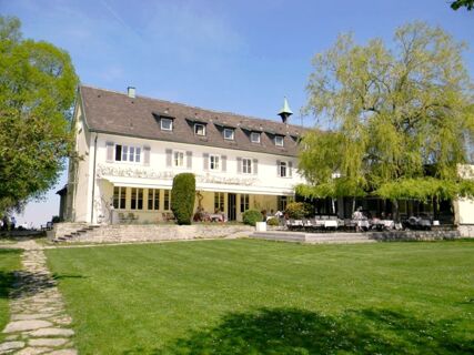 Das Hotel Landgut Burg zeigt eine wei&szlig;e Fassade mit mehreren Fenstern, einer Terrasse mit Sitzgelegenheiten und einer gro&szlig;en gr&uuml;nen Wiese im Vordergrund unter blauem Himmel.