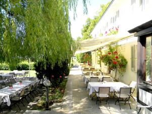 Au&szlig;enbereich mit bestuhlter Terrasse und wei&szlig;en Tischdecken neben dem Geb&auml;ude vom Hotel Landgut Burg, umgeben von gr&uuml;nen Pflanzen und B&auml;umen.