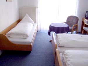 Hell eingerichtetes Hotelzimmer mit Einzelbett, Doppelbett, Tisch und Stuhl im Gasthof Schwarzer Adler mit Blick auf einen Vorhang.
