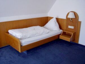 Schlichtes Einzelzimmer mit Holzbettrahmen und Nachttisch, beleuchtet von einer Wandlampe im Gasthof Schwarzer Adler.